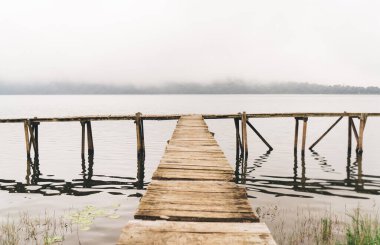 Sis ve kasvetli gökyüzü ile kaplı sakin göl kenarında uzun tahta iskele manzarası.
