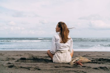 Genç, hayalperest bir kadının, yaz yolculuğunda esintinin tadını çıkarırken, kum torbasında sallanan sicim torbasıyla oturuşunun arka görüntüsü.