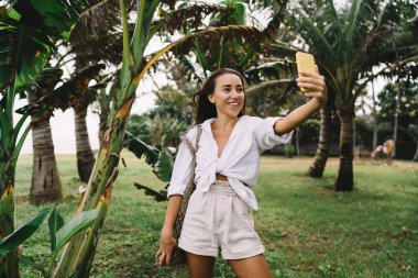 Pozitif genç bir kadın, tropikal parktaki yeşil ağaçların yanında tatilde palmiye ağaçlarıyla durup cep telefonuna bakıyor ve selfie çekiyor.