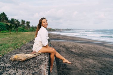 Bir çanta dolusu meyveyle gezen, kameraya bakıp gülümseyen ve tropik plajda serinleyen iyimser kadın yolcuların yan görüntüsü.