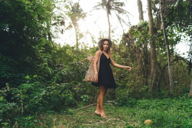 Hasır şapkalı genç kadın vücudu güneşli bir günde yeşil ormanda yürürken kameraya bakıyor ve tatilin tadını çıkarıyor.