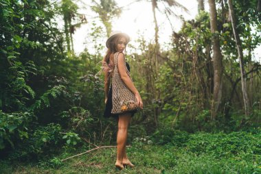 Yeşil ormanda duran ve yaz mevsiminde el çantasıyla kameraya bakan şapkalı genç bir kadın.