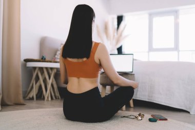 Siyah taytlı, turuncu bluzlu, Lotus pozlu, netbook kullanarak internette yoga yapan zayıf bir kadının arka planı.