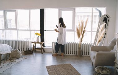 Sıradan giysiler içinde, cam pencerenin yanında cep telefonuyla dikilip evdeki rahat oturma odasında ekranı seyreden kimliği belirsiz genç bir kadın görüntüsü.