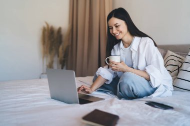 Genç, mutlu, siyah saçlı bir kadın rahat bir yatakta rahatlarken netbook 'ta sosyal ağlarda geziniyor ve bir bardak sıcak içki içiyor.