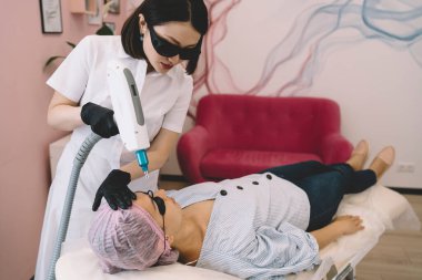 Koruyucu gözlüklü ve tek kullanımlık şapka takan kadın müşteri, profesyonel kadın terapist lazerle yüz kıllarını aldırma işlemi yaparken rahatlıyor.