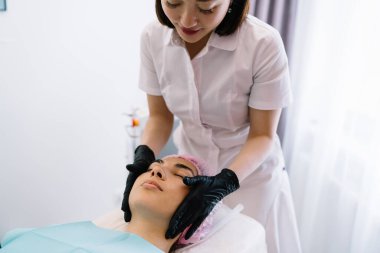 Beyaz üniformalı profesyonel. Güzellik salonunda yaşlanma karşıtı tedavi sırasında gözleri kapalı bir şekilde kanepede yatan kadının yüzüne masaj yapıyor.