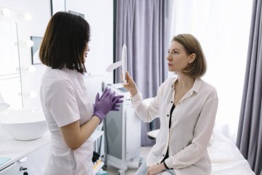 Aynada oturan ve güzellik kliniğindeki randevusu sırasında uzmanların tavsiyelerini dinleyen konsantre kadın hastaların yüksek açılı görüntüsü.