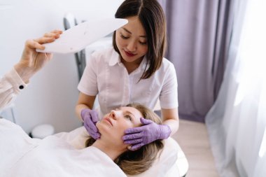 Kanepede uzanan ve modern ışık salonundaki güzellik prosedüründen sonra aynaya bakan yüksek açılı bir kadın hasta.