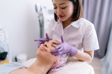 Ekin içeriği profesyonel Asyalı kadın güzellik uzmanı lateks eldivenler, ameliyat sırasında gözleri kapalı bir şekilde müşterinin yüzüne masaj yapıyor.