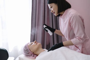 Uzman Asyalı güzellik uzmanı güzellik salonundaki masada yatan kadının yüzüne şişeden kozmetik sprey sıkar.