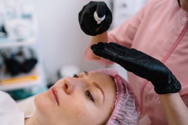 Koruyucu eldivenli isimsiz kadın kozmetikçi müşteriyle yüz bakımı ameliyatına hazırlanırken nemlendirici losyon sürüyor.