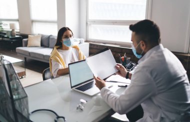 Koruyucu maskeli konsantre bir erkek doktor dizüstü bilgisayarla masada oturuyor ve yüzündeki kadın hasta raporlarını inceliyor.