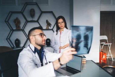 Masada dizüstü bilgisayarla oturan genç ve pozitif erkek doktor, modern klinikte doğal ışığa karşı film çekerken, film alanındaki kadın meslektaşlarını gösteriyor.