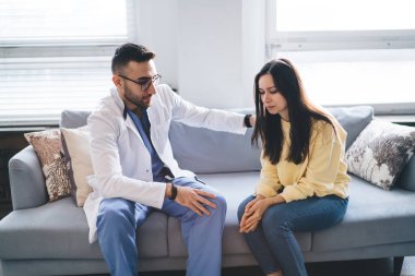 Özel üniformalı ve gözlüklü ciddi bir erkek onkolog koltukta otururken teşhis hakkında bilgi verdikten sonra dikkatle bayan müşterinin omzuna dokunuyor.