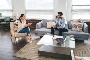 Günlük giysiler ve gözlüklerle ilgili erkek terapist kanepede oturuyor ve rahat ofisinde terapi sırasında kadın hastaların zihinsel sorunları hakkında konuşmalarını dinliyor.