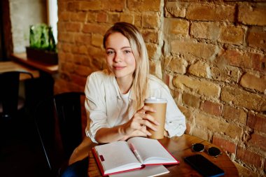 Modern kafede ders çalışmaktan bir fincan kahveyle rahatlarken kameraya bakıp gülümseyen kız öğrencilerden.