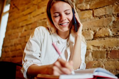 İyimser bayan girişimcinin cep telefonuyla konuşma yapması ve kafeden uzaktan çalışırken planlayıcıya bilgi yazması.