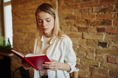 Ciddi bir serbest çalışan, sarı saçlı, günlük kıyafetli. Yaratıcı eğlence alanında tuğla duvarın yanında dururken not defterinden bilgi okuyor.