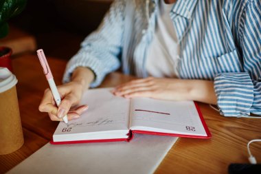 Kafede cep telefonuyla çevrimiçi derslere katılırken, elinde kahve fincanı ile ahşap masada oturan ve not defterine notlar yazan anonim bayan öğrencilerden.