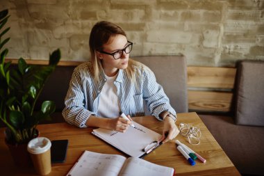 Modern kahve evinde çalışırken, günlük giysiler ve gözlüklerle serbest çalışan düşünceli kadın çizim şeması yüksek açılı.