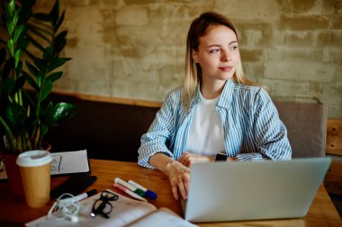 Akıllı saatli genç bir bayan, bilgisayar kırtasiyesi kahve fincanı ve akıllı telefonla masada otururken aynı zamanda yeni yazılım geliştirme projesine hizmet veriyor ve modern kafeye bakıyor.