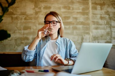 Akıllı saati olan, gözü kapalı bardaktan su içen ve ön planda laptopuyla masada oturan, kafeden uzaktan kumandalı yazılım projesi üzerinde çalışan serbest çalışan bir kadın.