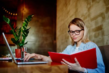Gözlüklü genç bir kız öğrenci elinde akıllı telefon kahve bardağıyla masada oturuyor ve kafede sınav hazırlıkları sırasında dizüstü bilgisayarında not tutuyordu.