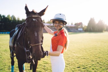 Kask ve üniformalı genç bir binici yaz aylarında çimenlik alanda bulutsuz gökyüzüne karşı antrenman yaparken kestane atının yanında duruyor ve kameraya bakıyor.