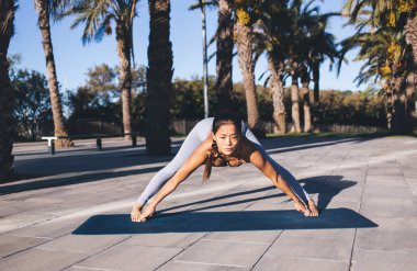 20 'li yaşlarda sıradan bir eşofman giymiş, sokakta palmiye ağaçlarıyla yoga antrenmanı yapan Asyalı bir kadın paspas üzerinde egzersiz yaparken vücut dengesini kontrol ediyor ve dışarıda esniyor.