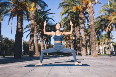Spor eşofman giymiş spor bir kadın sokakta palmiye ağaçlarıyla bütünsel iyileşme için yoga yapıyor, Asyalı kadın paspas üzerinde egzersiz yaparken vücut esnekliğini kontrol ediyor.