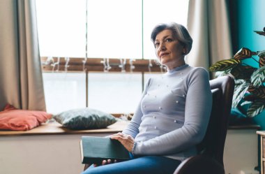 Düşünceli orta yaşlı kadın pencerenin yanında elinde kitapla otururken ve saksı bitkileriyle odada vakit geçirirken başka tarafa bakıyor.