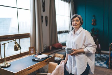 Kendine güveni tam orta yaşlı bir kadın şık giyiniyor masanın yanında duruyor ve kameraya bakarken elinde bir fincan sıcak içecek tutuyor.