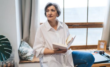Düşünceli orta yaşlı kadın, gündelik giysiler içinde pencere eşiğinde oturuyor ve açık bir kitap tutuyor.