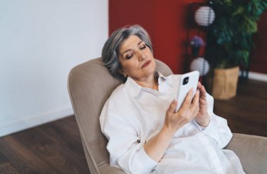 Yüksek açılı, şık giyinen, rahat bir oturma odasında dinlenirken koltukta oturup akıllı telefonlara bakan olgun bir kadın.