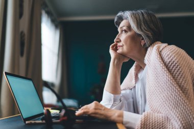 Düşünceli kadın serbest çalışanın, gündelik kıyafetlerle masada dizüstü bilgisayarla oturması ve proje üzerinde çalışırken gözlerini kaçırması.