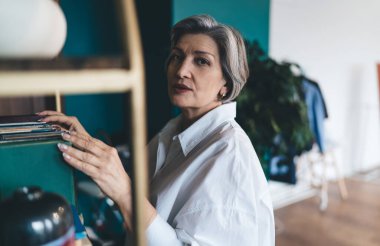 Kendinden emin orta yaşlı bir kadın, elleri kitapların arasında dururken kameraya bakıyor ve bulanık bitkilerle dolu bir odada zaman geçiriyor.