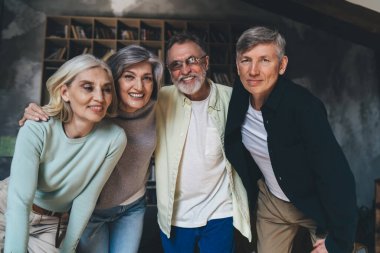 Dost canlısı, orta yaşlı, gündelik giysiler içinde birbirine yakın duran ve modern çatı katı stüdyosundaki kameraya bakan insanların toplu portresi.