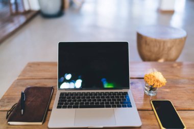 Netbook cep telefonundan oluşan modern iş yeri ve cam kavanozdaki sarı çiçeğin yanındaki ahşap masaya yerleştirilmiş kalemli not defteri.