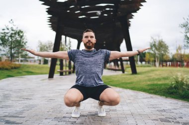 Spor salonunda spor yaparken kollarını kaldırıma uzatarak çömelmiş çömelme hareketleri yapan ciddi sakallı bir erkek sporcu.