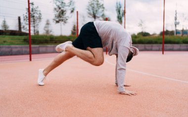 Aktif giyimli, metal çitlerin ve bulutlu gökyüzünün altındaki ağaçların yakınındaki stadyumda amuda kalkma egzersizi yapan isimsiz sporcu.