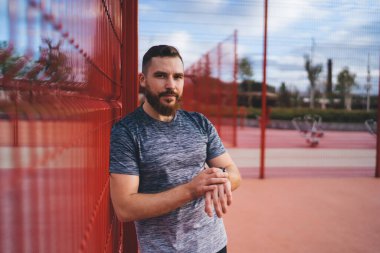 Spor salonundaki genç sakallı adam el ele tutuşup kırmızı metal çitlere ve bulanık ağaçlara bakarken kol saati takıyor.