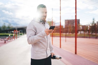 Spor kıyafetleriyle gezen genç sporcu yaz günü spor sahasında spor yaparken cep telefonu kullanıyor.