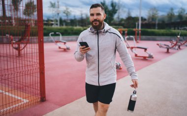 Spor kıyafetleri içindeki sakallı erkek sporcu ve bir şişe su kameraya bakarken metal çitlerin yanında spor sahasında yürüyordu.