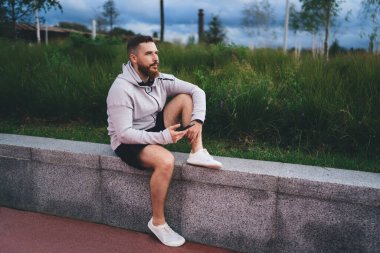 Sakallı erkek sporcu taş bir bankta oturuyor ve parka bakarken cep telefonu kullanıyor.