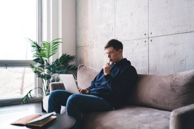 Genç serbest çalışan düşünceli bir erkek bilgisayar ekranına bakarken ve evde boş zamanlarında uzak bir proje üzerinde çalışırken kanepede oturuyor.