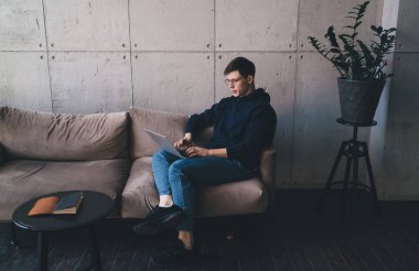 Rahat giyinen ve gözlüklü yüksek açılı bir adam dizüstü bilgisayarını kullanarak koltukta oturuyor ve evde uzak bir proje üzerinde çalışıyor.