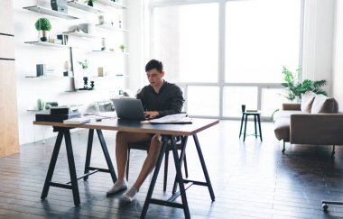 Portatif bilgisayar kullanan ve geniş oturma odasındaki masada otururken görüntülü konuşma yapan tüm erkek serbest çalışan.
