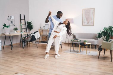 Neşeli Afro-Amerikan ruh eşleri günlük kıyafetleriyle birbirlerine bakıyorlar ve birlikte salonda eğlenerek dans ediyorlar.