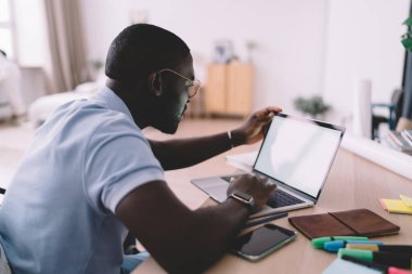 Sıradan kıyafetler ve gözlüklerle evde dizüstü bilgisayarla çalışan Afrikalı Amerikalı bir erkeğin yan görüntüsü.